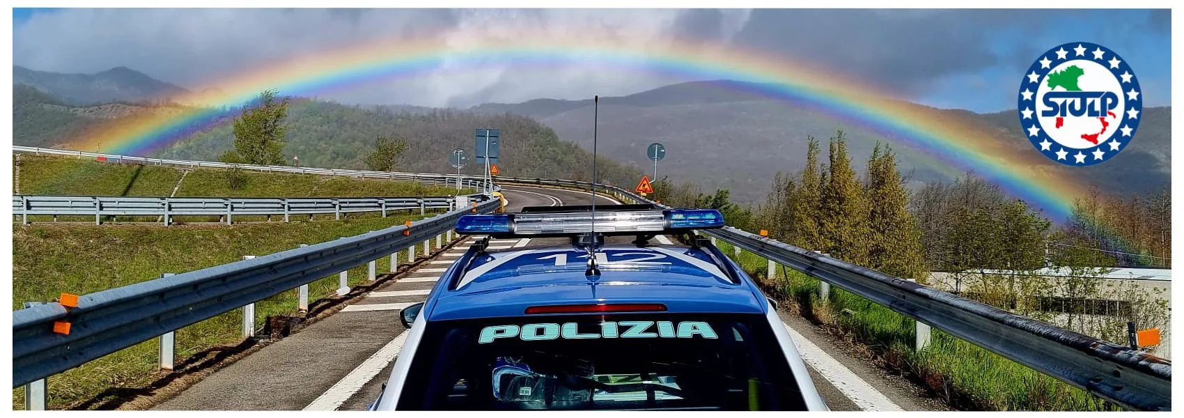 Un'auto della Polizia Stradale su una strada di montagna, con un arcobaleno sullo sfondo, presso il passo di Berceto.